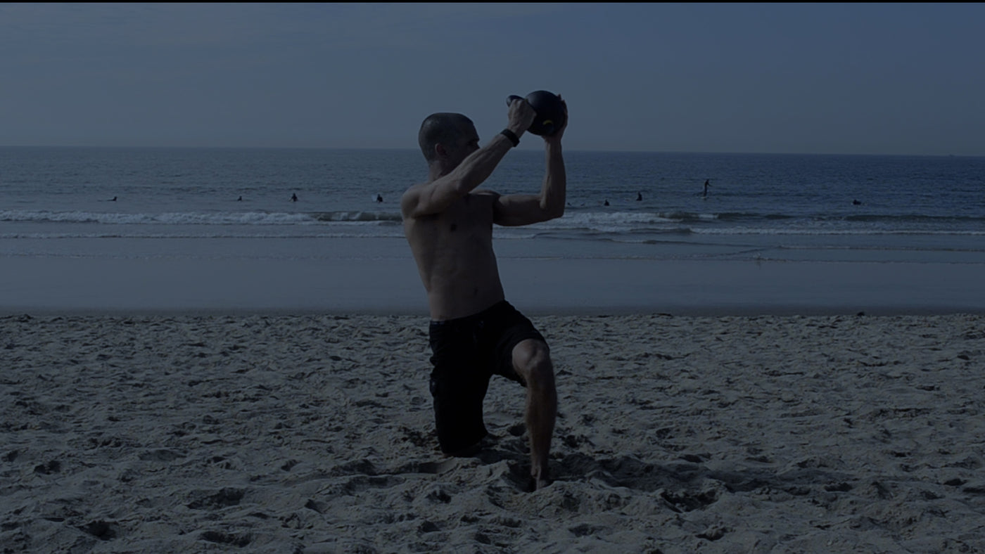 Man performing a half kneeling dynamic diagonal lift with a kettlebell on the beach