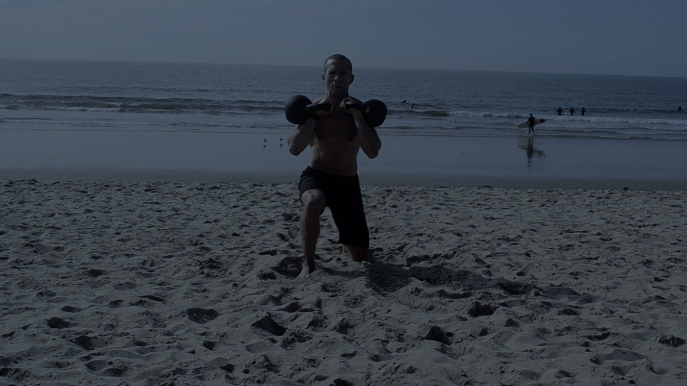 Man performing a racked double kettlebell reverse lunge on the beach